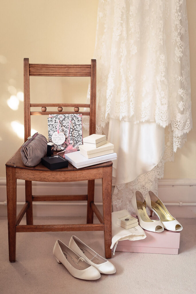 A wooden chair holds stacked books and accessories, with wedding shoes arranged on the floor nearby against a lace curtain backdrop.