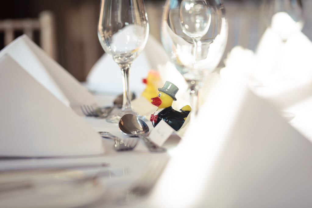 A formal place setting with wine glasses and a small yellow rubber duck wearing a top hat and bow tie as a table decoration.