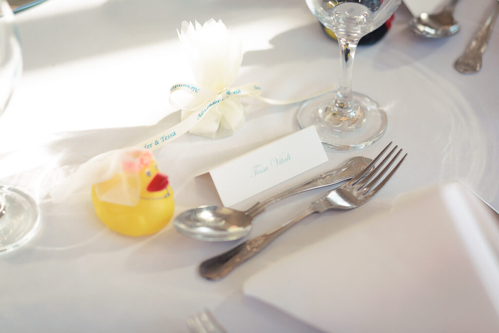 A formal place setting with a white napkin, cutlery, wine glass, and a yellow rubber duck as a whimsical decoration.