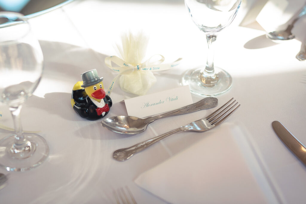 A formal place setting with a decorative rubber duck wearing a top hat, place card, silver cutlery, and water glass on white tablecloth.