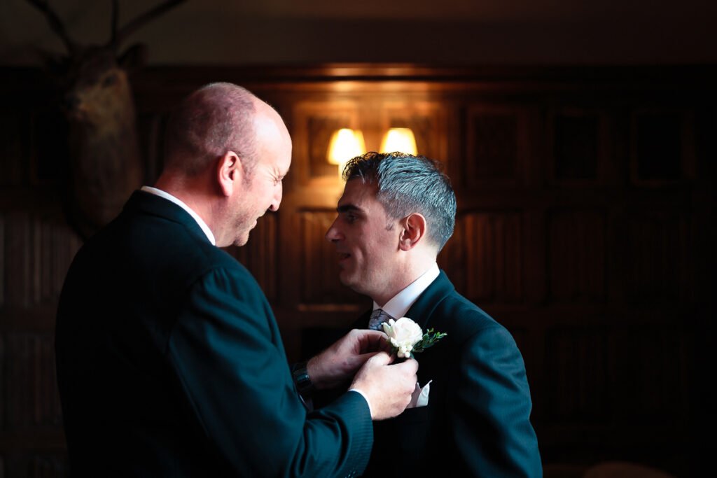Two men in dark suits face each other indoors. One adjusts a white flower boutonniere on the other's jacket. Warm lighting behind them.
