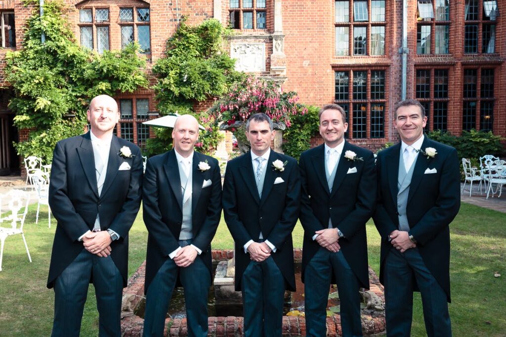 Five men in formal wedding attire stand in a row in a brick courtyard, wearing dark suits and buttonholes.