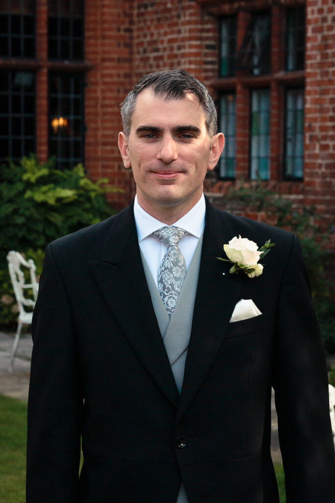 A man in formal attire with a boutonnière stands in front of a brick building at what appears to be a wedding event.