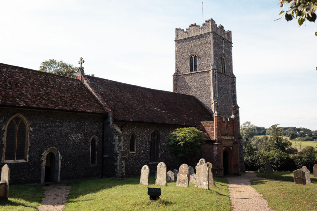 A stone church with a brick tower stands in a graveyard with headstones, surrounded by green fields and trees under a clear sky.