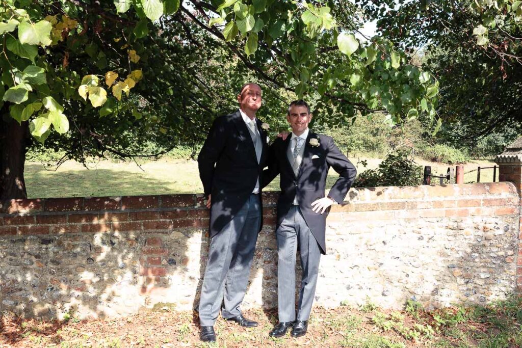 Two men in formal suits stand together against a brick wall beneath overhanging tree branches on a sunny day.