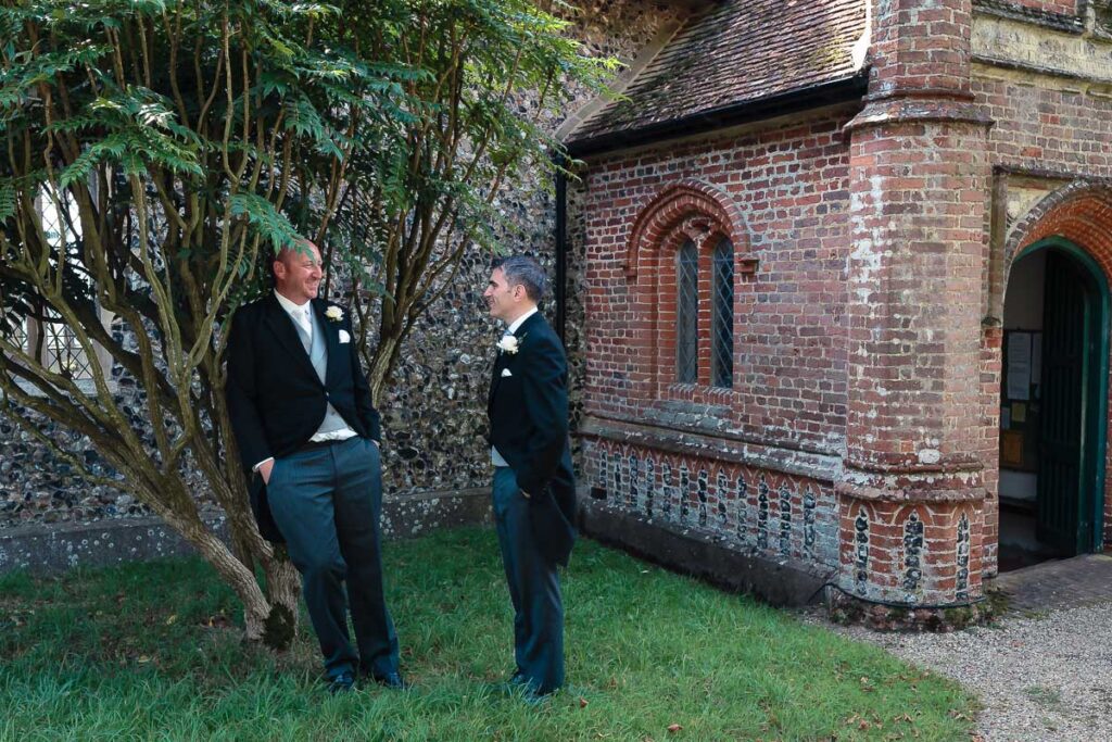 Two men in dark suits with white buttonholes stand talking beside a gnarled tree near a red brick building with a green door.