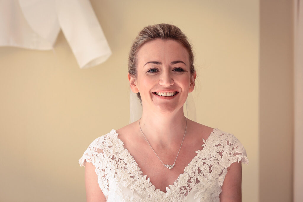 A woman in a white lace dress and necklace smiles at the camera in a beige interior setting.