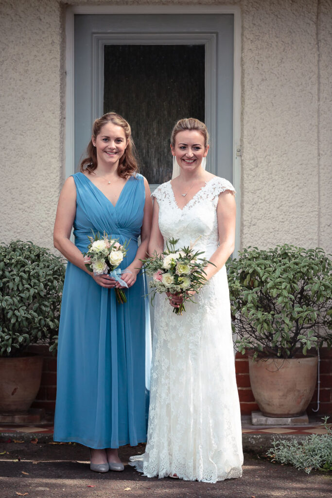 Two women in formal attire stand smiling before a doorway, holding bouquets of flowers.