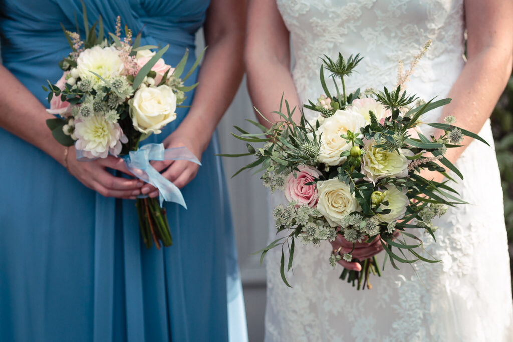 Two women in wedding attire holding bouquets of flowers. One wears a blue dress, the other a white lace gown.