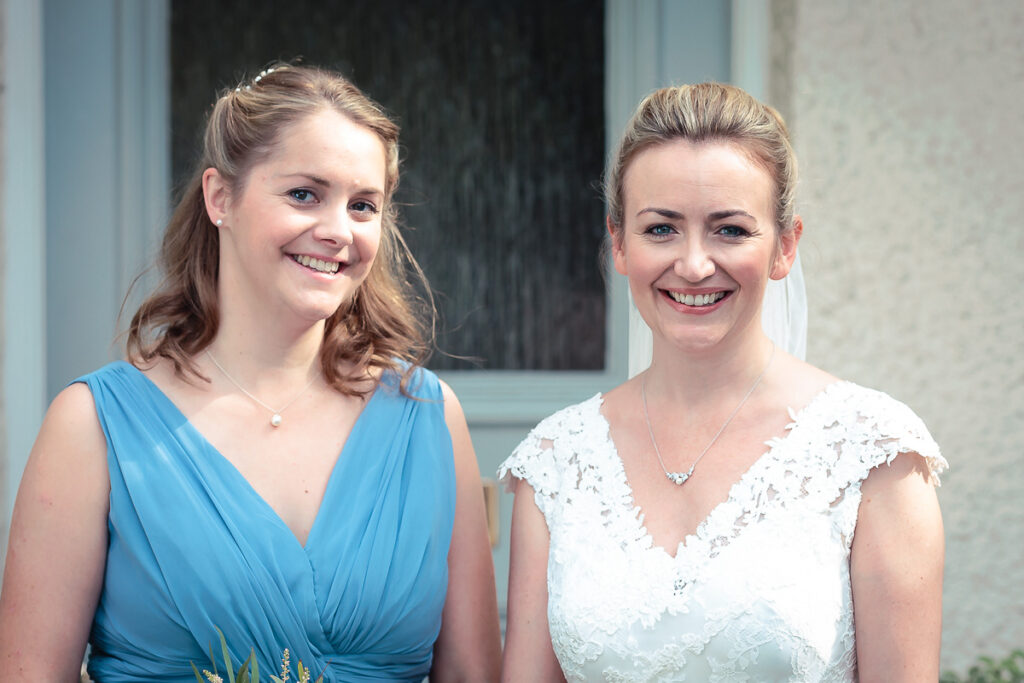 Two young women smiling at the camera outdoors. One wears a blue dress, the other a white lace dress. Classical building backdrop.