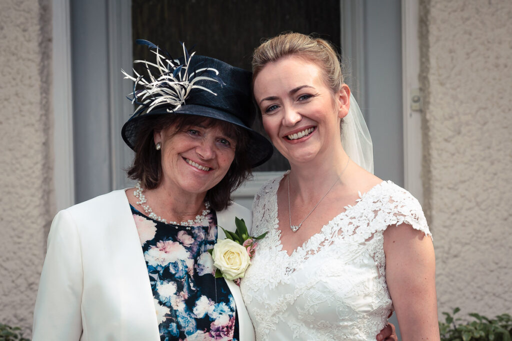 Two women smiling at the camera outside a building. The woman on the right wears a white lace wedding dress and veil. The woman on the left wears a cream jacket, floral dress, and decorative hat.