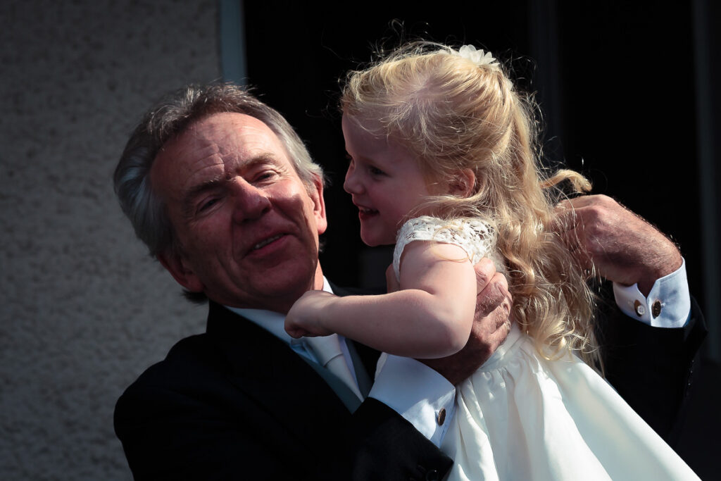 An older man in a dark suit holding a young girl in a white dress, both smiling, against a dark background.
