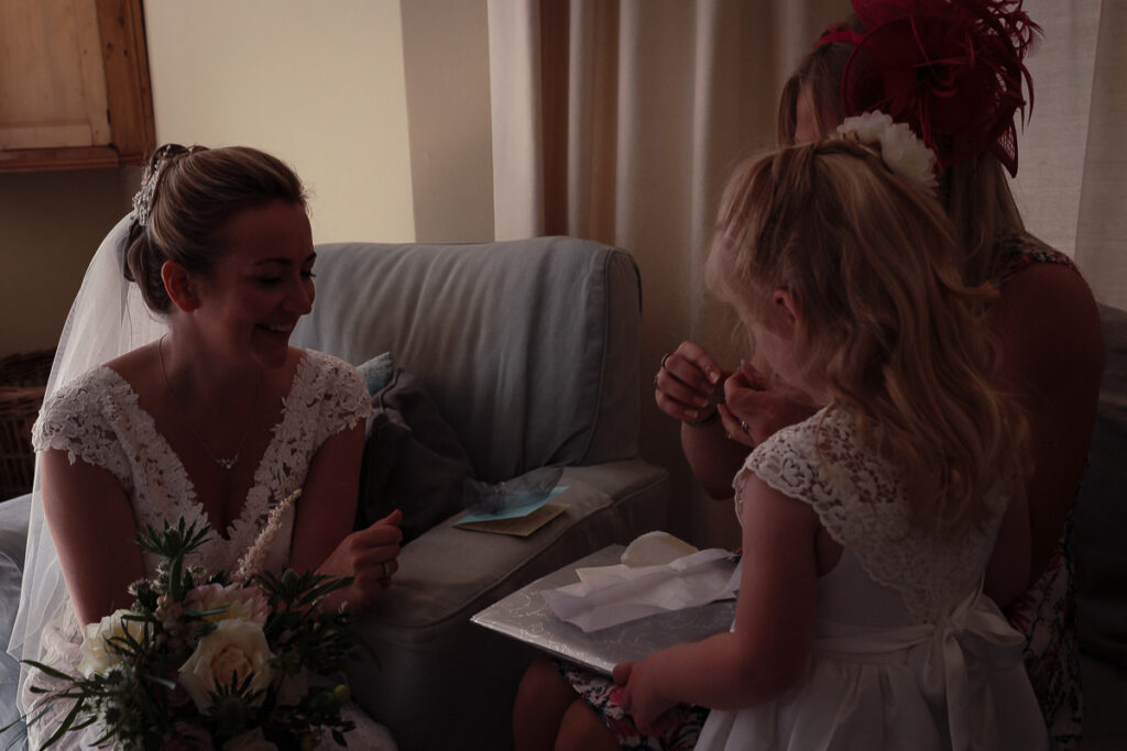 Two women in white lace dresses sit on a grey sofa, smiling and conversing. One wears a veil and holds a bouquet. Papers lie on the sofa between them.