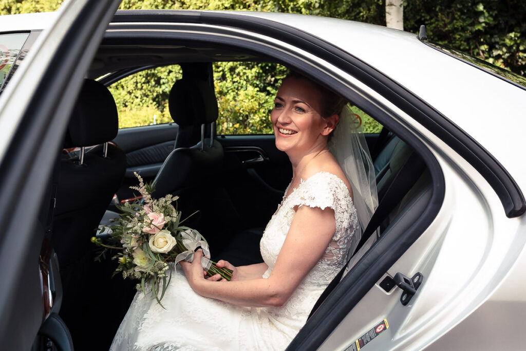 A bride in a white lace dress sits in the back seat of a white car, holding her bouquet and smiling at the camera.