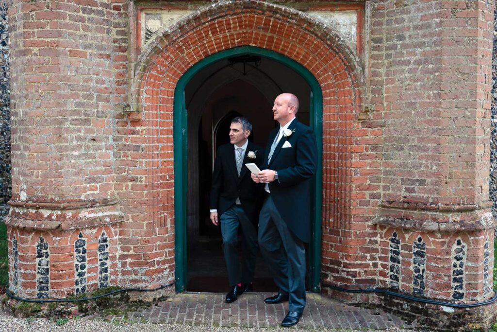 Two men in formal wedding attire exit through a large arched brick doorway with a green frame.