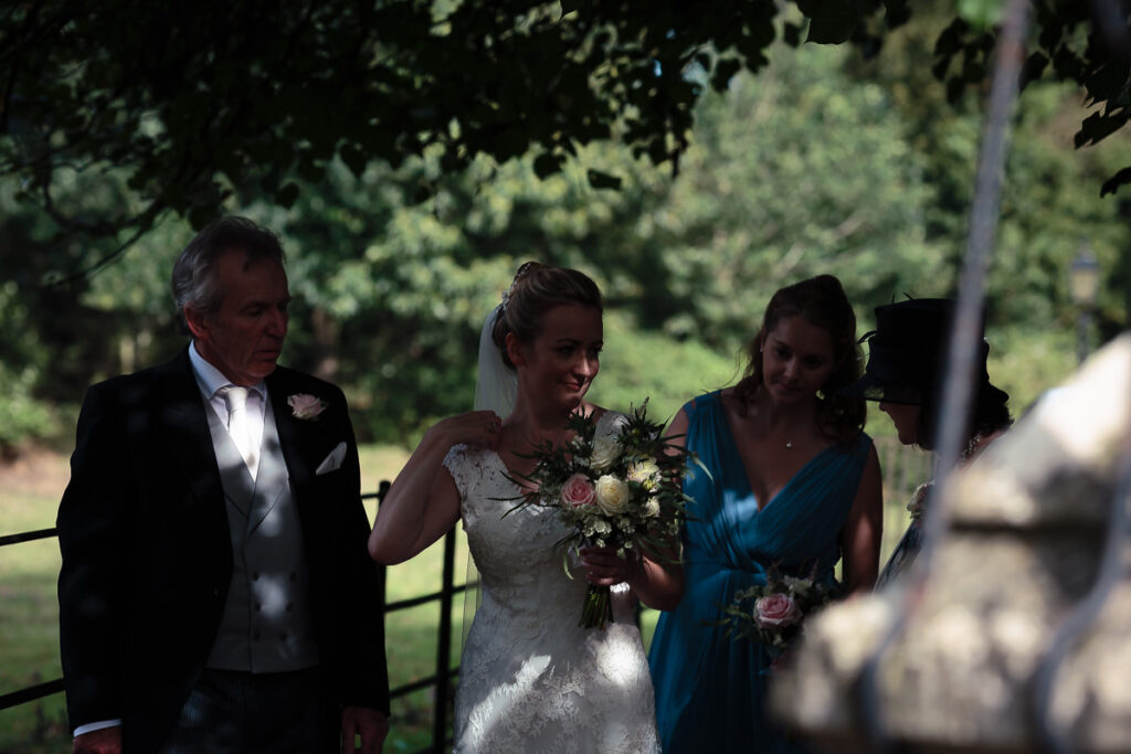 A bride in a white lace dress holds a bouquet while standing with an older man in a tuxedo and a woman in a blue dress outdoors among trees.