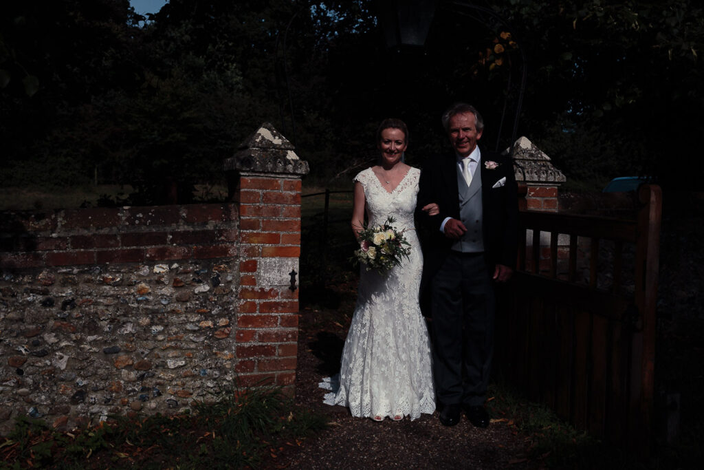 A bride in a white lace dress and groom in a grey suit stand together by an old brick gateway, holding a bouquet.