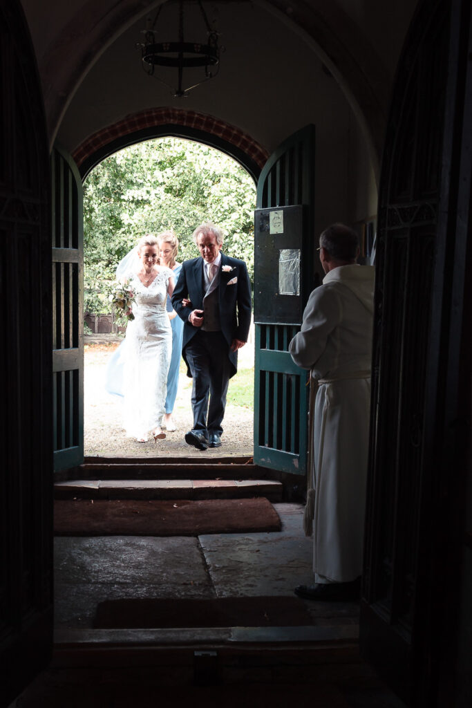 A bride and groom walk through an arched stone doorway towards a garden, with a clergy member in robes standing in the foreground.