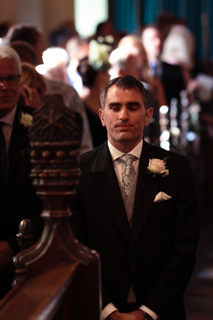 A man in a black suit and patterned tie with a boutonnière stands in a church aisle, looking downward during what appears to be a wedding ceremony.