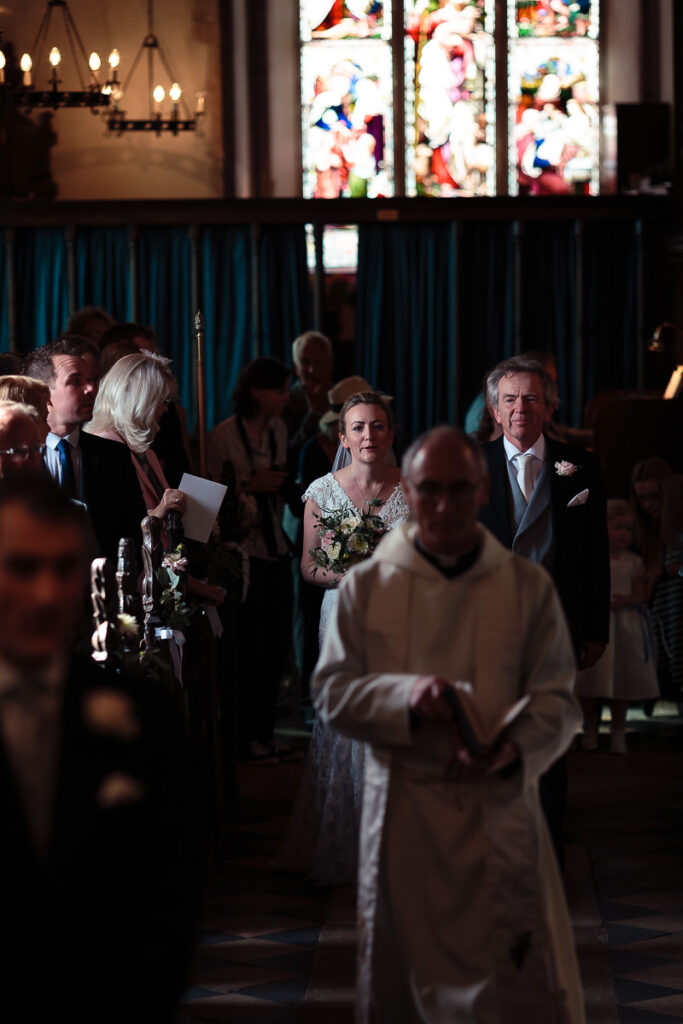 A wedding ceremony in a church. A bride in a white dress holds flowers whilst standing beside a groom in a suit, with clergy and guests present.
