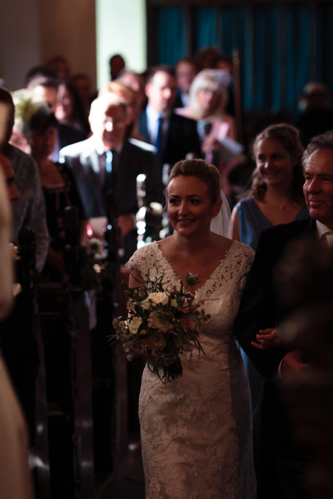 A bride in a lace wedding dress holds a bouquet, smiling among guests at an indoor wedding ceremony.