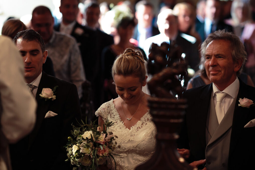 A bride in a white lace dress walks with two men in formal attire during a wedding ceremony, holding a bouquet of flowers.