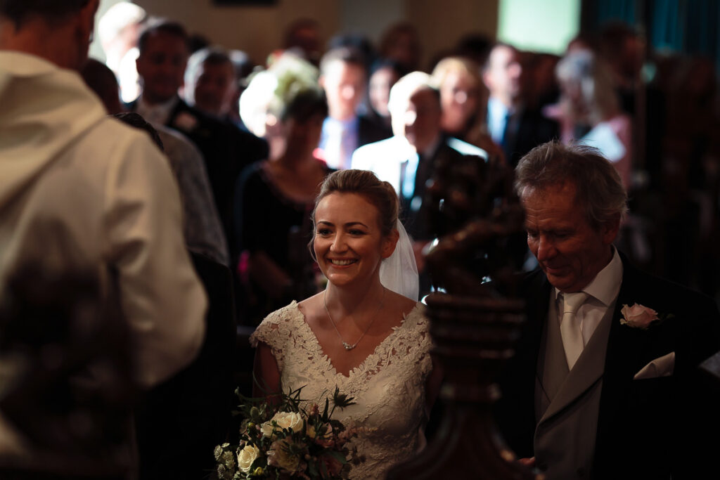 A bride in a lace wedding dress and veil smiles while walking with an older man in a suit at a wedding ceremony, surrounded by guests.