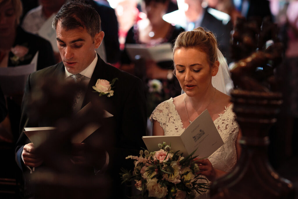 A bride and groom stand together during their wedding ceremony, both holding service booklets and looking down as they recite vows.