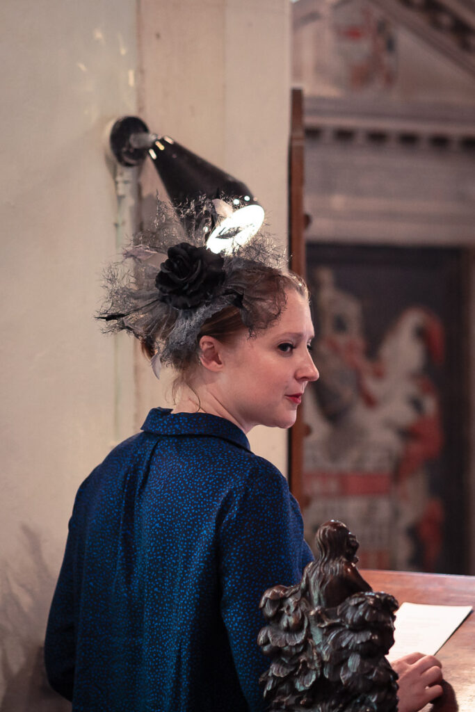 A woman in a blue jacket wears an elaborate black fascinator with netting and a decorative bird brooch on her head.