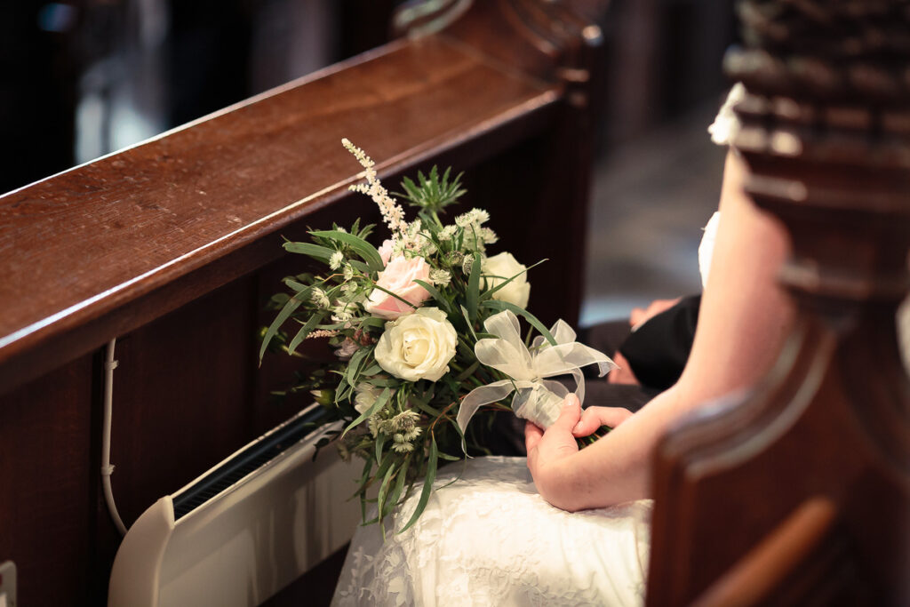 A bride holds a white rose bouquet tied with a sheer ribbon inside a wooden church pew during a wedding ceremony.