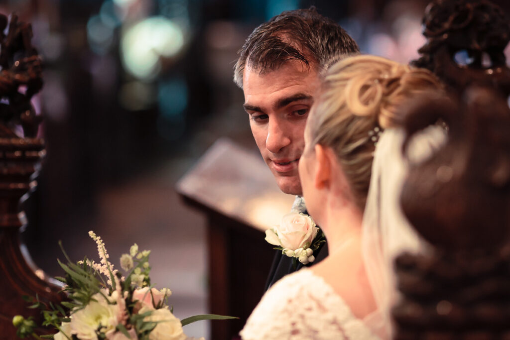 A man in formal attire smiles while speaking with a woman at what appears to be a wedding ceremony, with flowers visible.