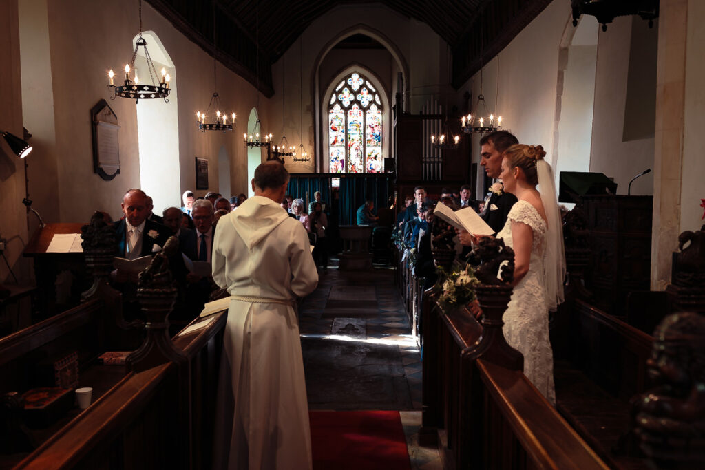 A wedding ceremony in a church. A bride in a white lace dress stands at the altar with a groom in a cream jacket, facing guests in wooden pews.