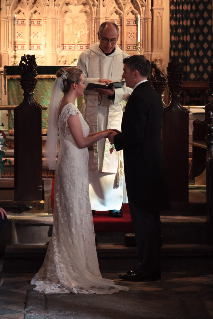 A wedding ceremony in a Gothic church. A priest in white robes holds a book between a bride in a lace gown and groom in a dark suit.
