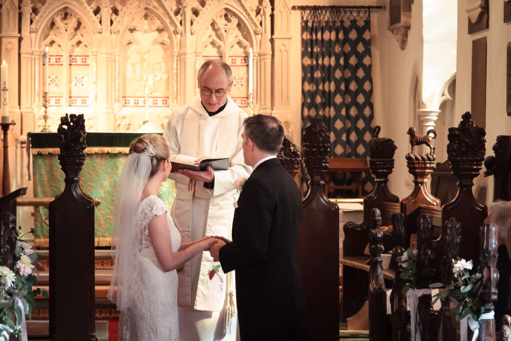 A wedding ceremony in a Gothic church. A priest in white robes conducts vows between a bride in a white lace dress and veil, and a groom in a black suit.