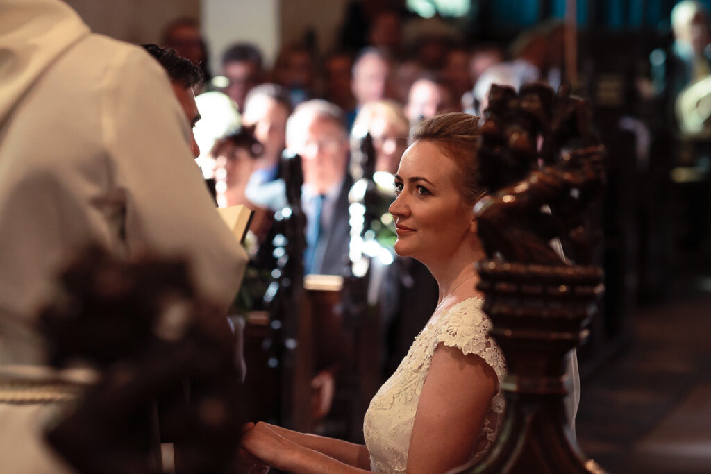 A woman in a white lace dress stands in profile at what appears to be a formal indoor event, with blurred guests visible in the background.