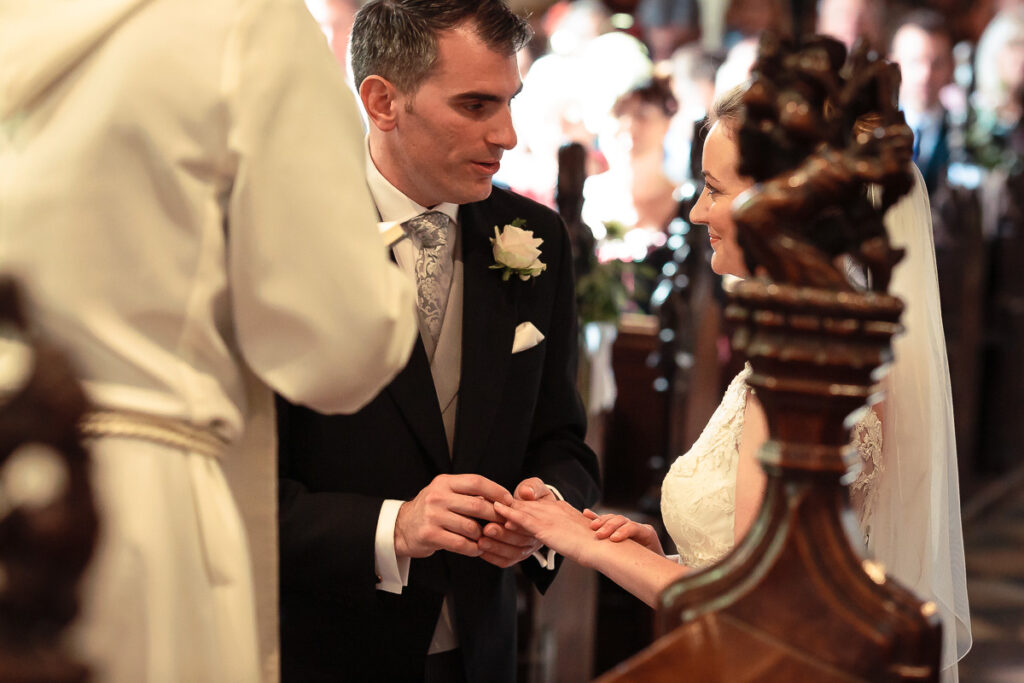 A groom in a black suit places a ring on the bride's finger during a wedding ceremony in a church.