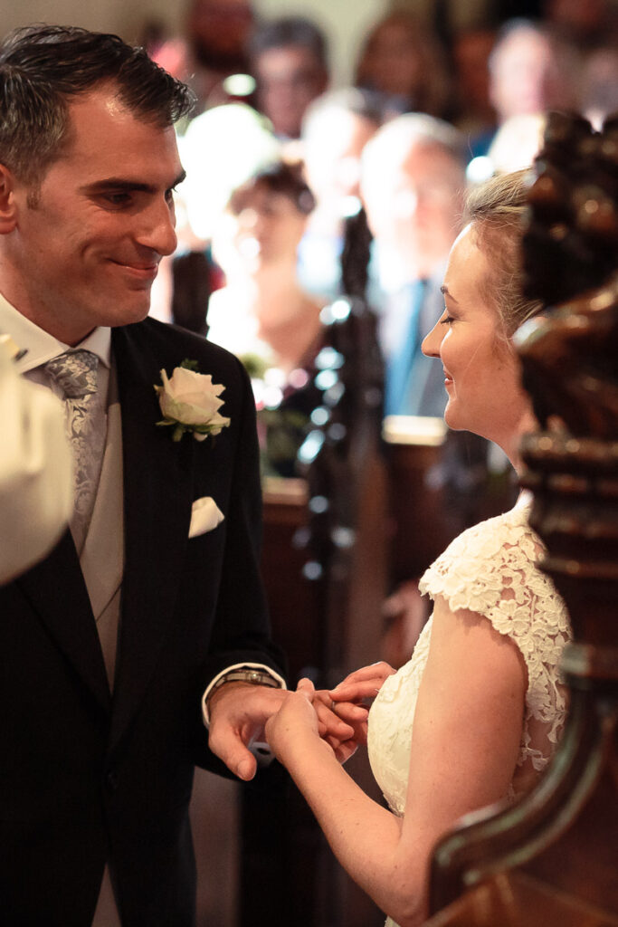 A couple exchanging rings during a wedding ceremony, with guests visible in the blurred background.