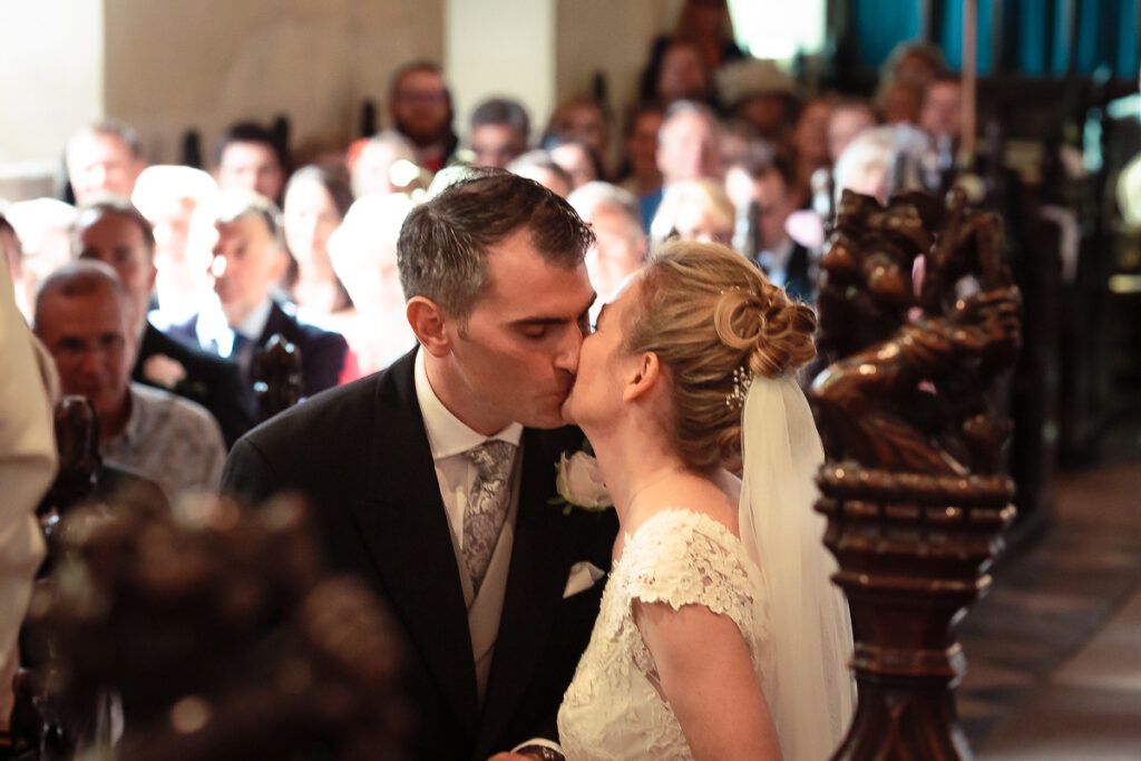 A bride and groom kiss at the altar during their wedding ceremony, with guests gathered behind them in a church.