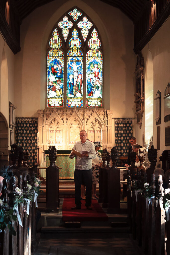 A man reads at a church altar beneath a tall stained glass window, flanked by dark wooden pews decorated with white flowers.