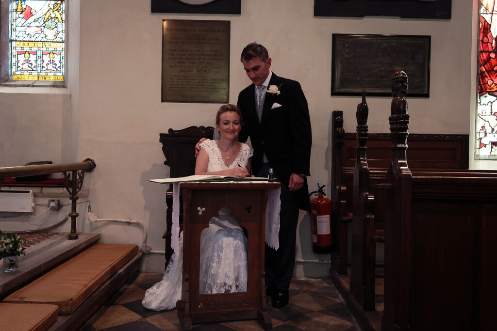 A bride in a white lace dress sits at a wooden lectern in a church whilst a groom in a dark suit stands beside her during a wedding ceremony.