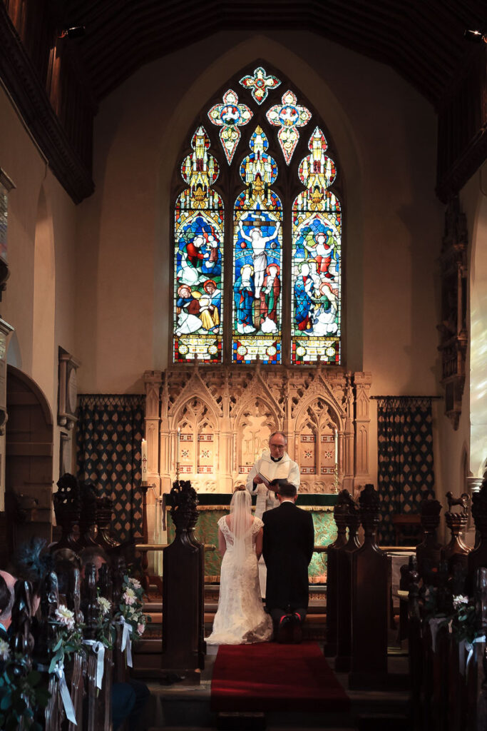 A wedding ceremony in a church, with a bride and groom at the altar beneath a large stained glass window.