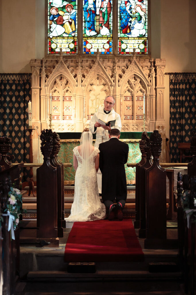 Wedding ceremony in a church. A bride and groom stand before an officiating clergyman at the altar, beneath stained glass windows.