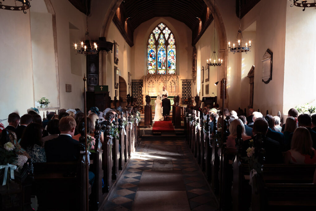 A wedding ceremony in progress inside a church, with guests seated in pews on either side of a central aisle leading to the altar.