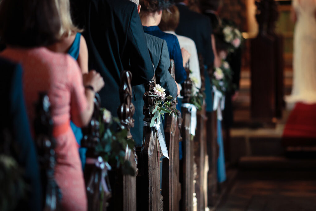 Church wedding ceremony with guests seated in pews decorated with white flowers and greenery along the aisle.