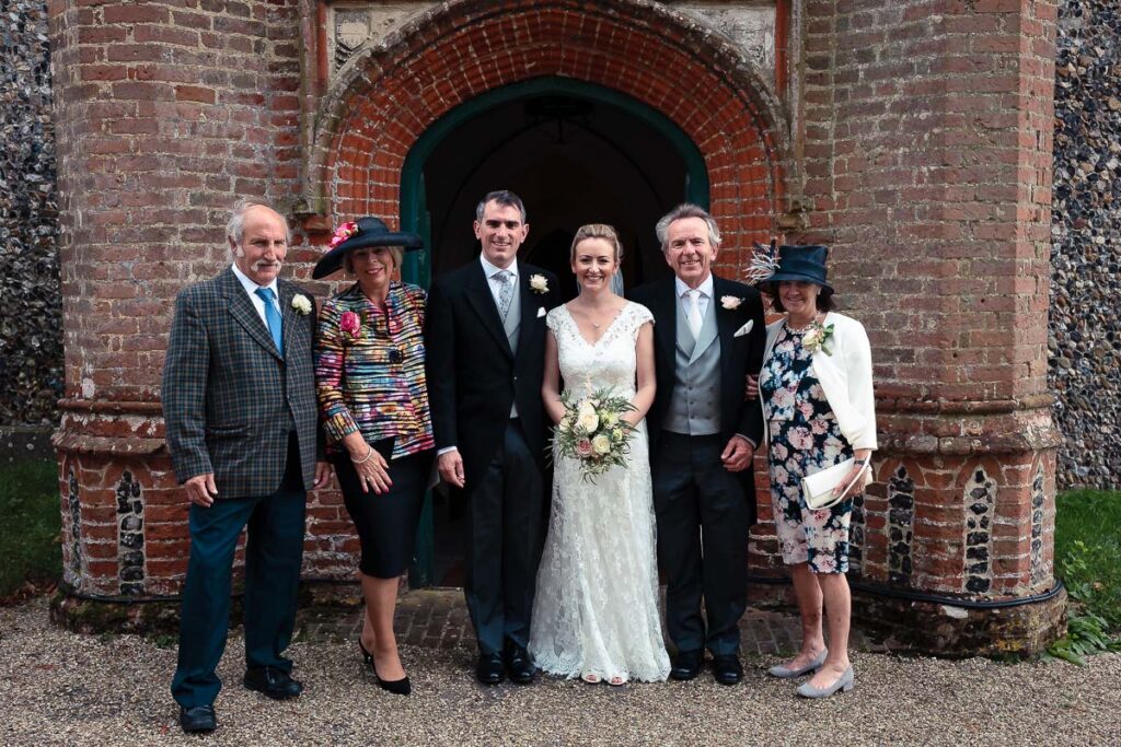 A bride and groom stand with four family members in formal attire outside a brick archway entrance.