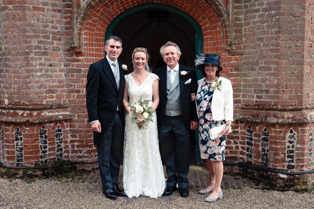 A bride in a white lace dress and groom with two older guests stand smiling before a red brick church archway.