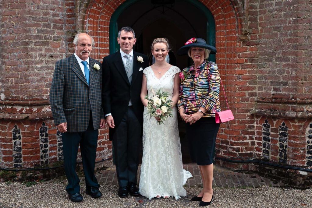 Four people stand outside a brick church entrance. A bride in a white lace dress holds a bouquet, flanked by a groom and two older guests in formal attire.