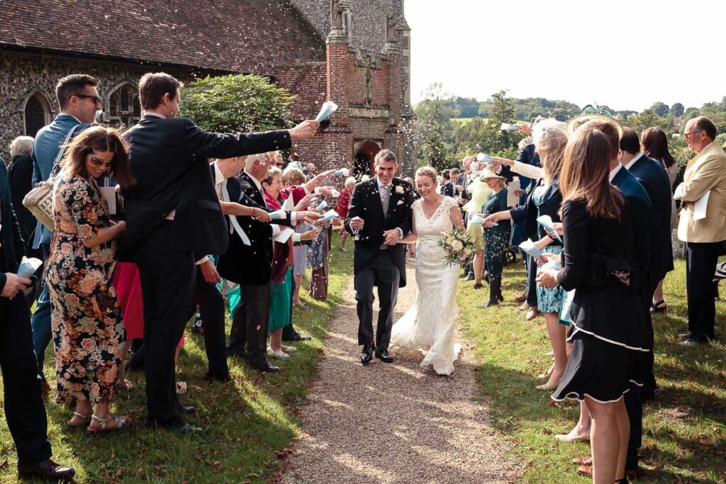 A bride and groom walk down a path between two lines of guests throwing confetti outside a brick church building.