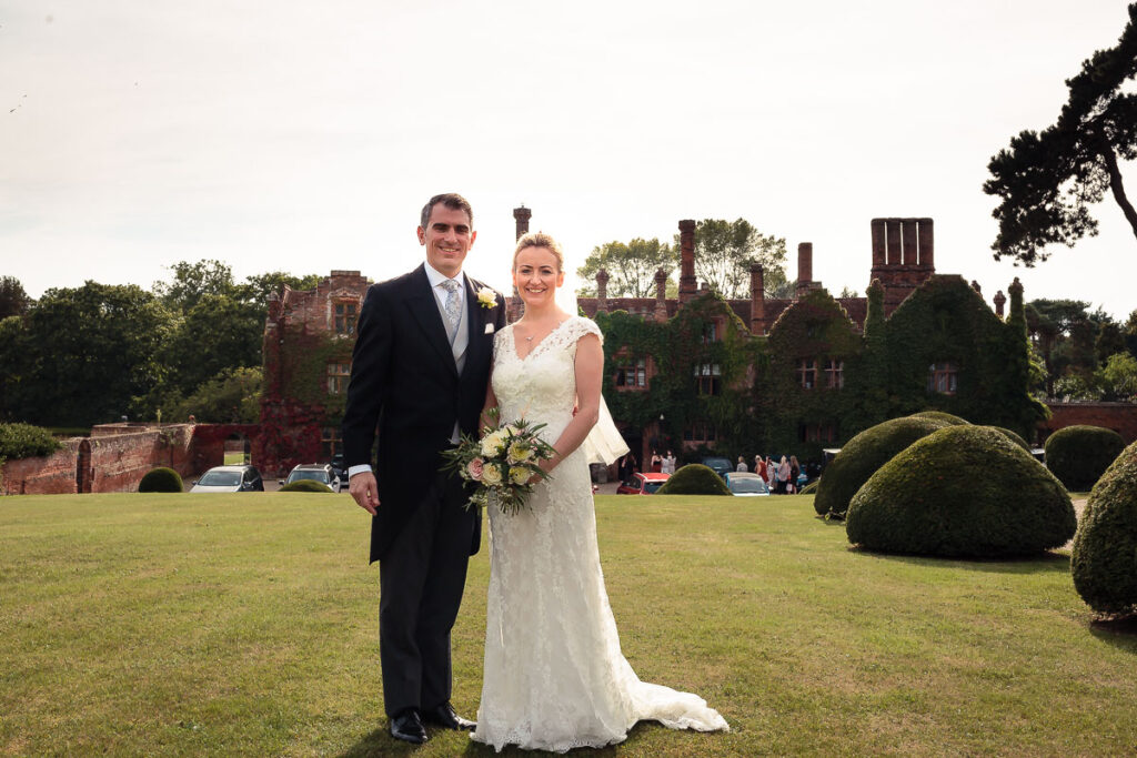 A bride and groom stand on a lawn before a historic brick mansion, smiling at the camera on their wedding day.