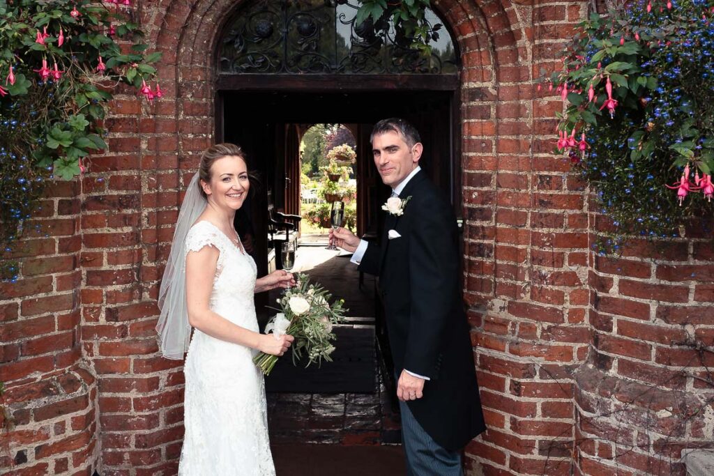 A bride and groom stand in a brick archway decorated with pink and blue flowering plants, smiling at the camera on their wedding day.